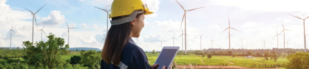 A woman engineer in a hard hat looks at an iPad with wind turbines in the background