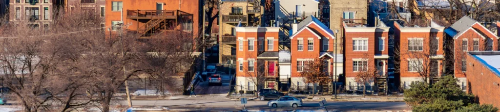 A row of red brick townhouses with white trim is situated along a street. In the foreground, there are leafless trees and patches of snow on the ground, while parked cars line the road.