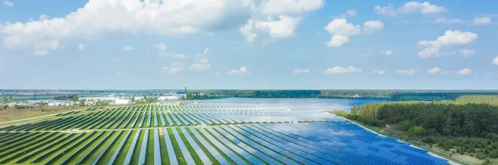 A large solar farm with rows of solar panels stretches across open fields under a partly cloudy sky, surrounded by greenery and distant buildings.